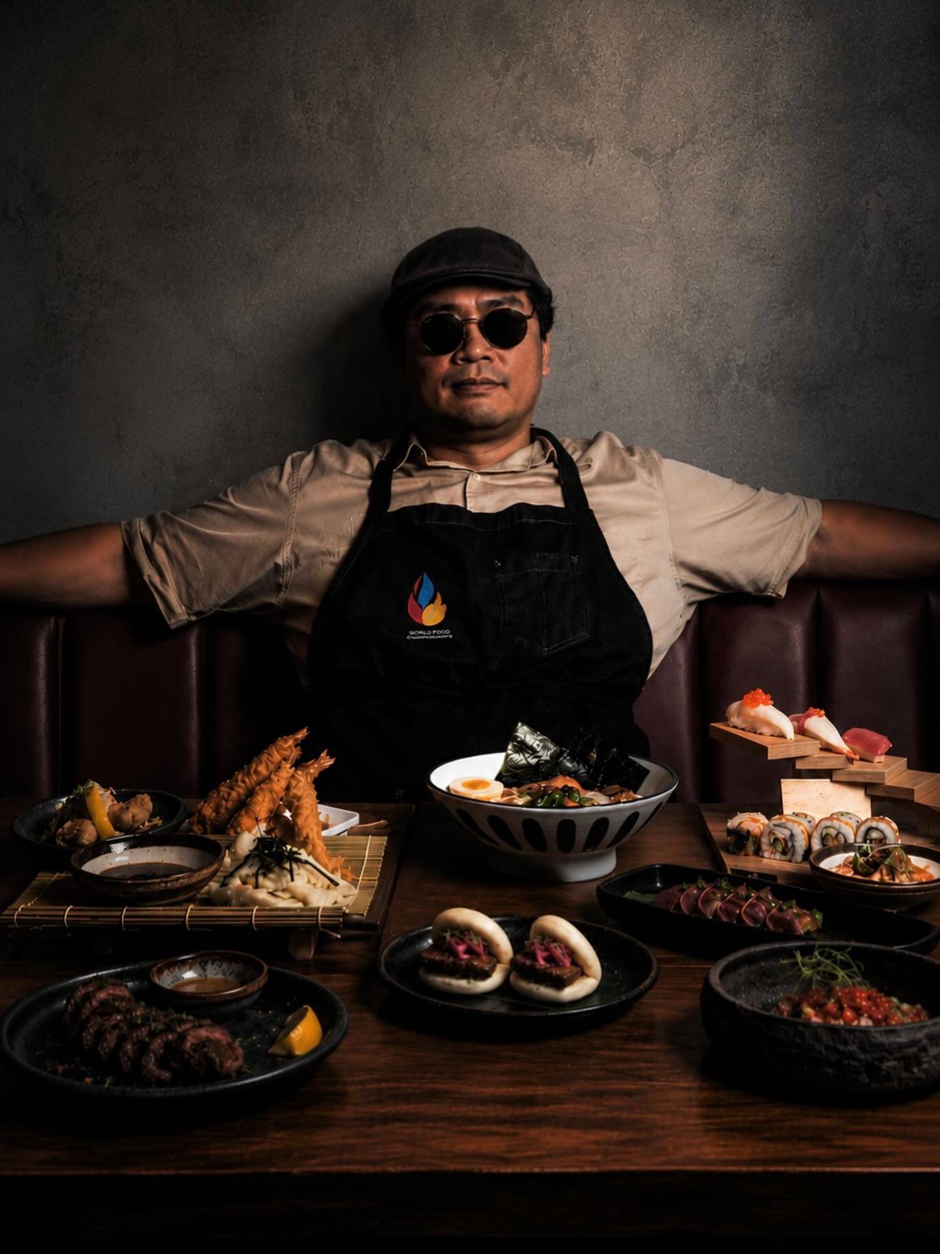 A chef in a black apron and sunglasses seated behind a table of sushi, nigiri, tempura and bowls, lit against a dark room.