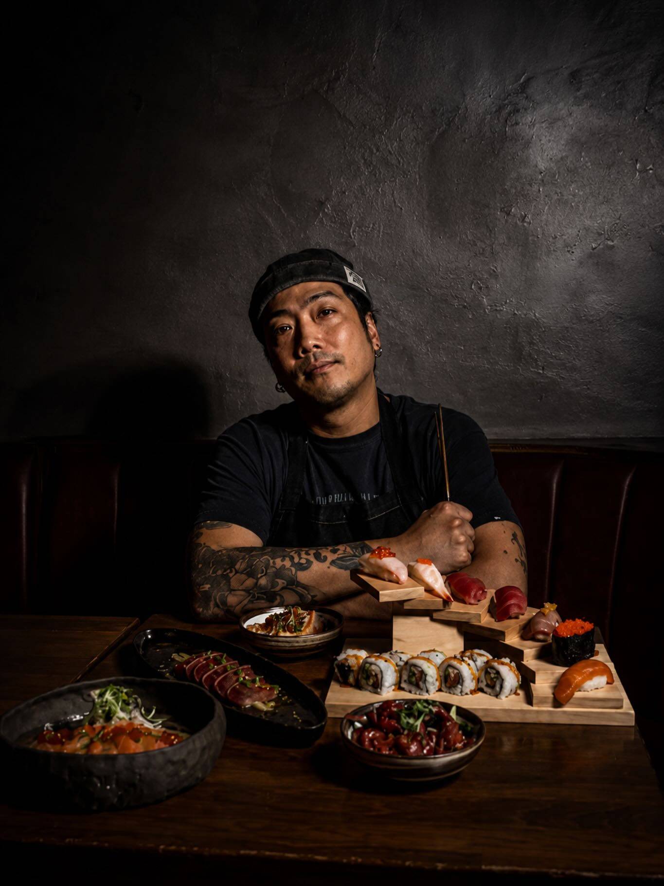 A chef with tattooed arms crossed, standing behind a spread of sushi and small plates in low light.